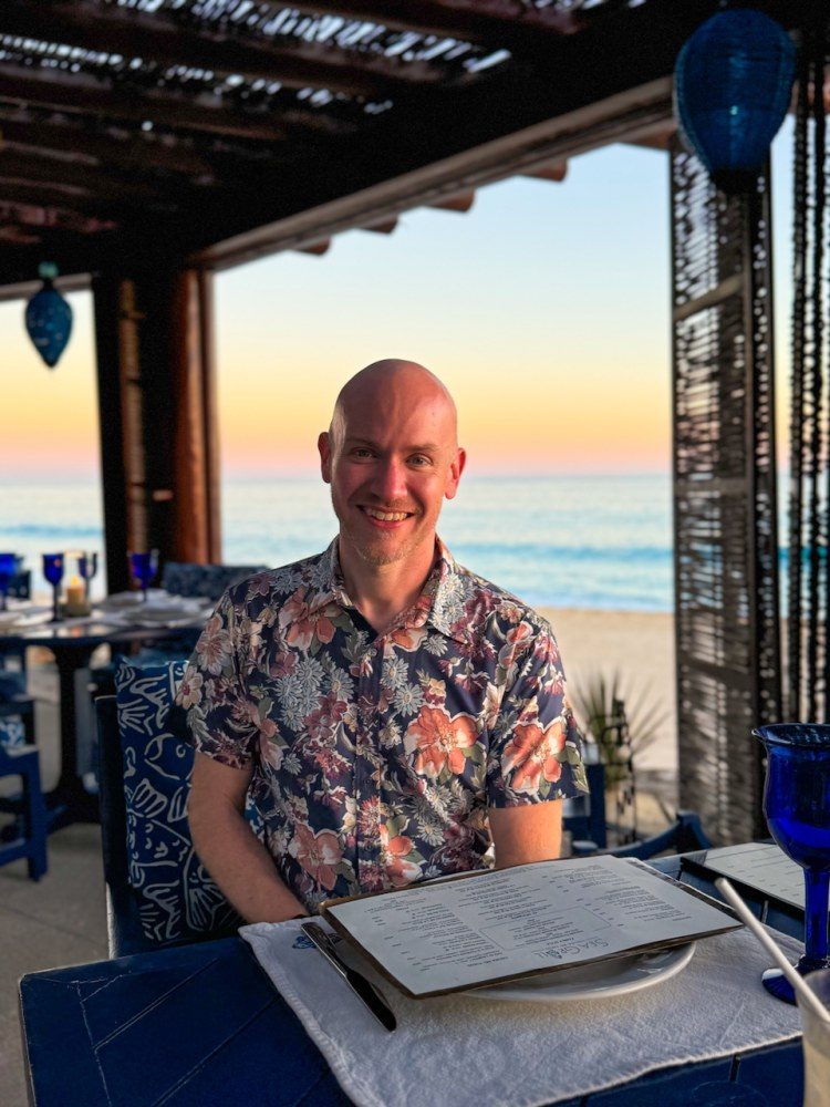 Jon Nickel-D’Andrea at a table by the sea at sunset