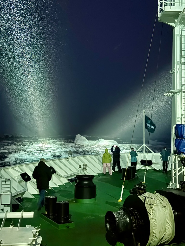 Passengers on the ship's bow at night, a searchlight beam full of snowflakes lighting pack ice and an iceberg ahead