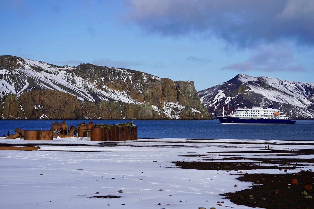Rusted whaling tanks on a snowy black-sand beach, an expedition ship at anchor below snow-streaked cliffs