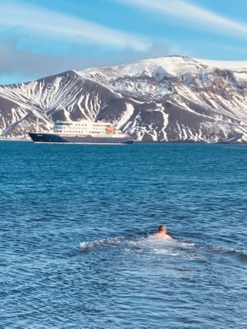 A swimmer in the dark blue water of the caldera, the Hondius at anchor below snow-covered volcanic slopes