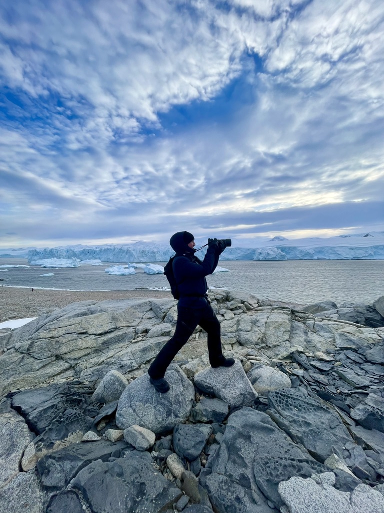 Claudio in a dark hooded jacket on grey rocks with his camera raised, an ice cliff and icebergs across the water behind
