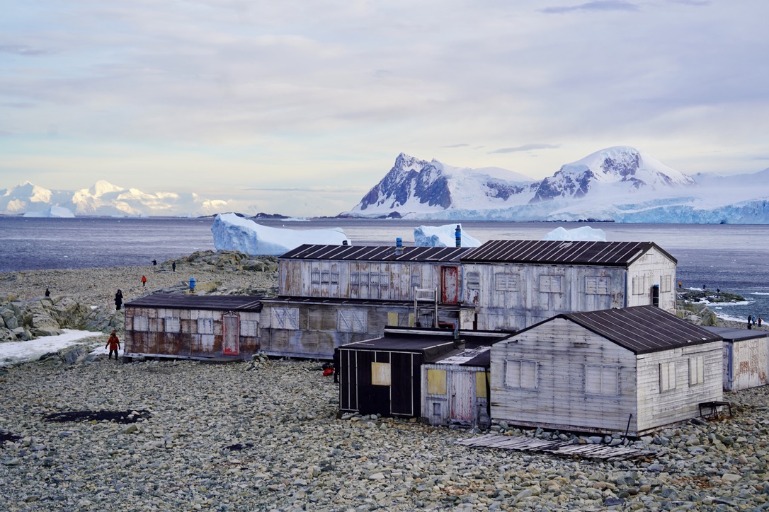 Weathered wooden huts of an abandoned research station on a rocky island, icebergs and snowy mountains behind