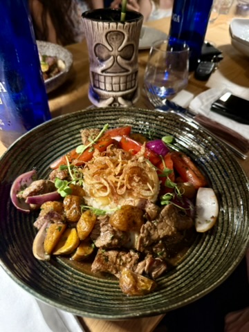 Braised meat with roasted vegetables and crispy onions in a dark bowl, a tiki mug behind it