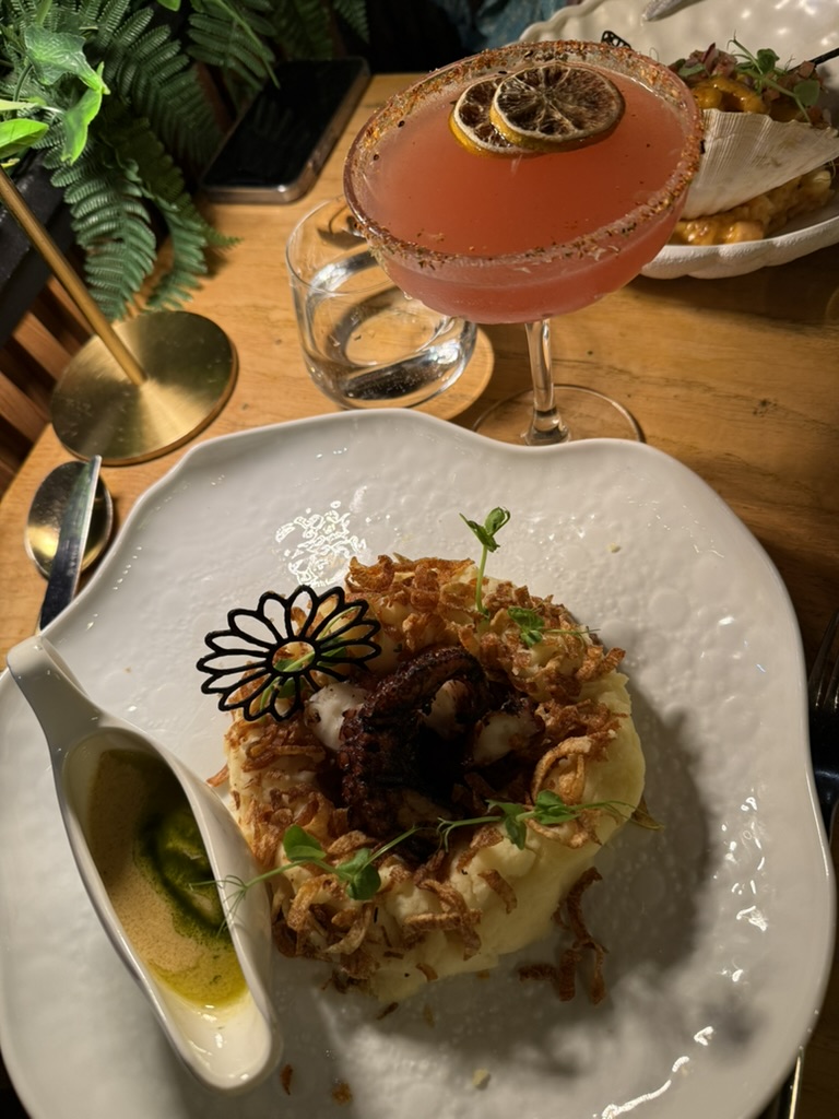 Grilled octopus on mashed potato with crispy onions, a pink cocktail behind it