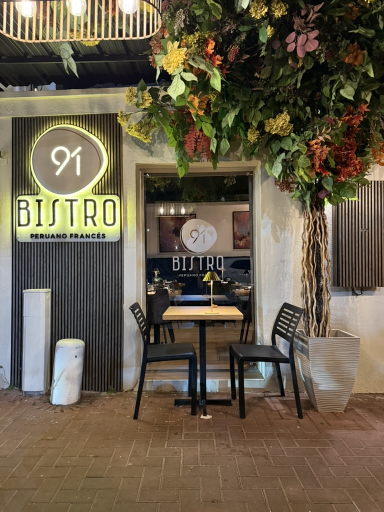 The lit sign of Bistro 91, Peruano Francés, beside a table for two under a flowering tree
