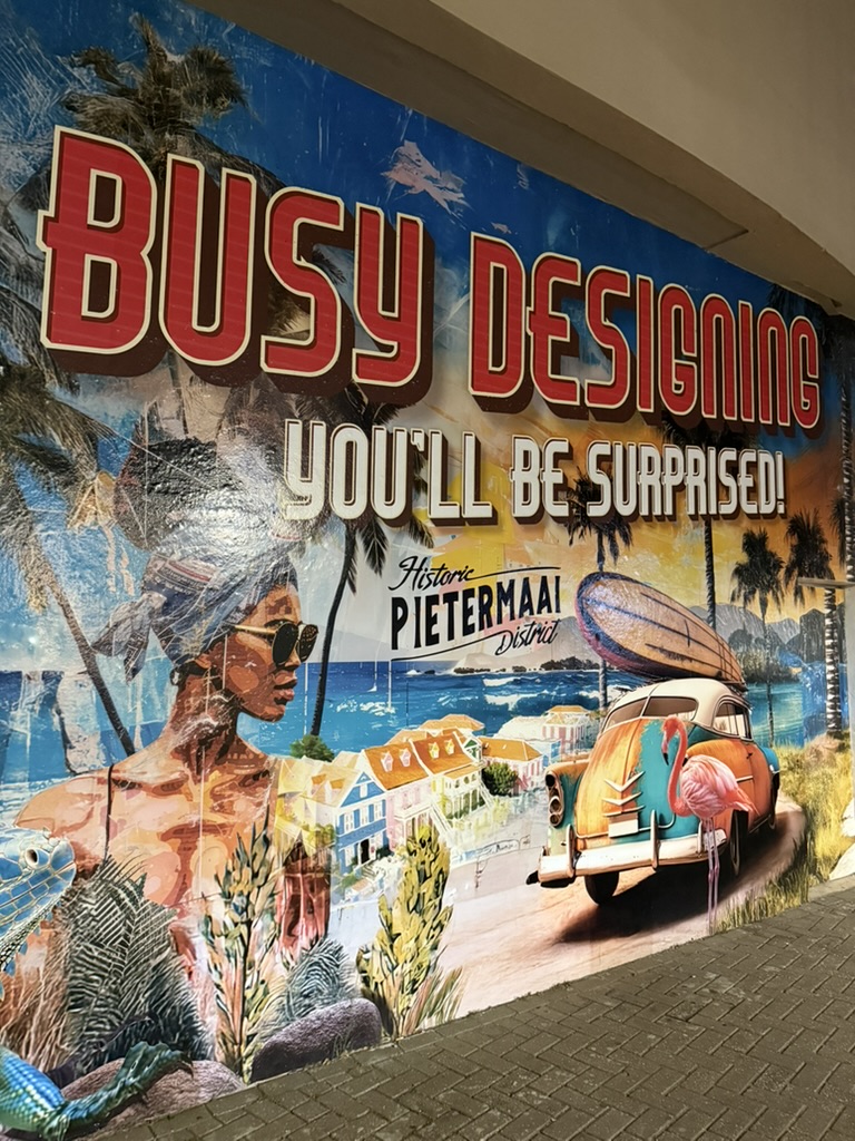 A large mural reading Busy designing, you'll be surprised, Historic Pietermaai District, with palm trees, a vintage car and a flamingo