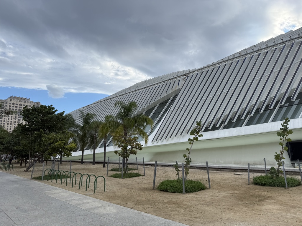 The long white side of the Museum of Tomorrow, lined with angled fins, palm trees in front under a cloudy sky