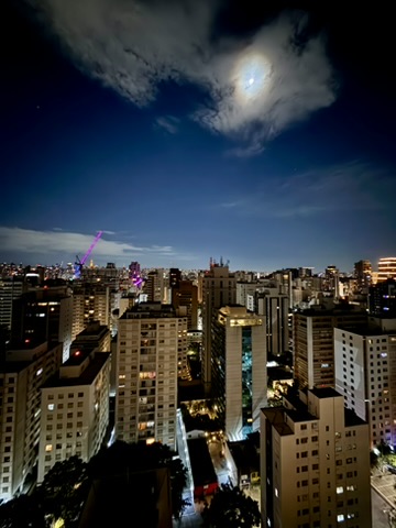 Night view over São Paulo's towers from a rooftop, a purple-lit construction crane, the moon behind clouds