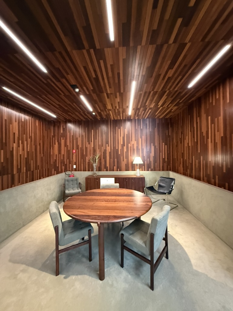A showroom clad in strips of dark hardwood on walls and ceiling, a round wooden table with tufted chairs, a black leather sling chair in the corner
