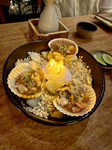 Three scallops in their shells on a bed of white salt, a flame in the middle of the dish, a wooden box with sake behind