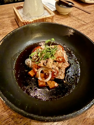 Slices of fish with cubes of sweet potato, red onion and microgreens in a dark glossy sauce on a black plate