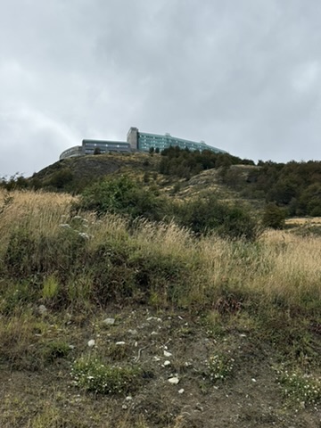 A long, low hotel of glass and stone along the top of a cliff, seen from the grassy slope below under a grey sky