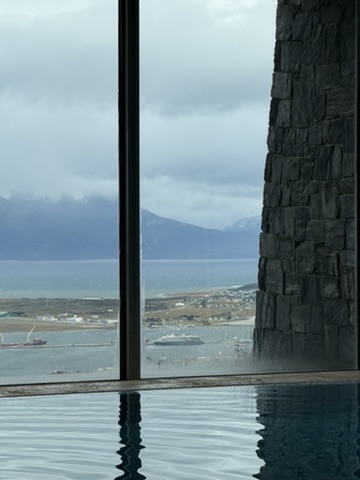 From inside the pool: a floor-to-ceiling window and a slate wall frame the view down over Ushuaia's airport peninsula, a cruise ship at the pier and the Beagle Channel