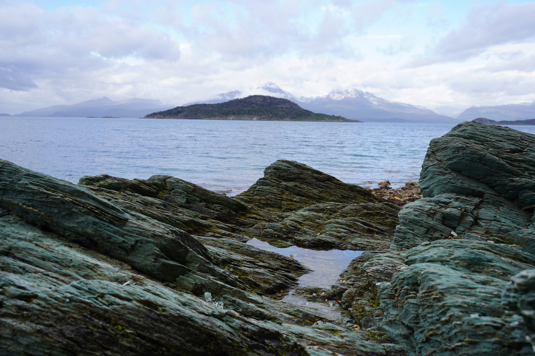 Green-grey layered rocks at the shore of the Beagle Channel, an island and snow-capped mountains across the water