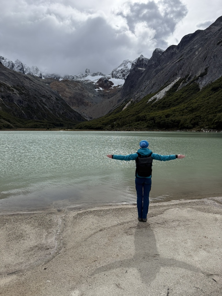 A hiker in a blue jacket stands on the sandy shore with arms spread wide, facing the turquoise lake and the snowy mountains