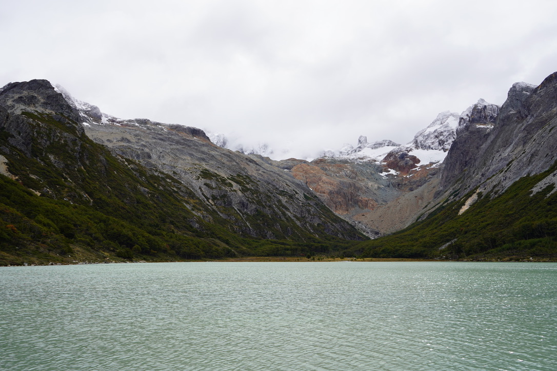 The full width of the lake, pale green water below dark forested slopes and a wall of grey and white peaks